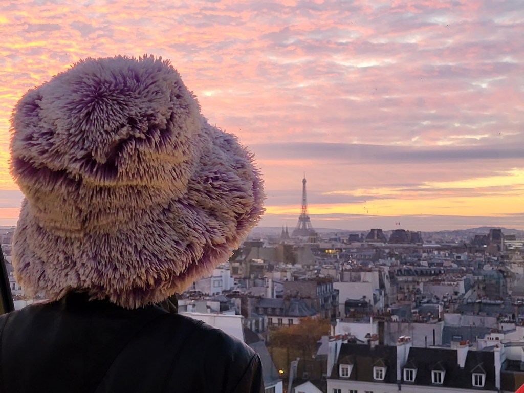 En haut de l&rsquo;escalator du musée Beaubourg, une voyageuse au beau chapeau face à la ville au&nbsp;crépuscule.