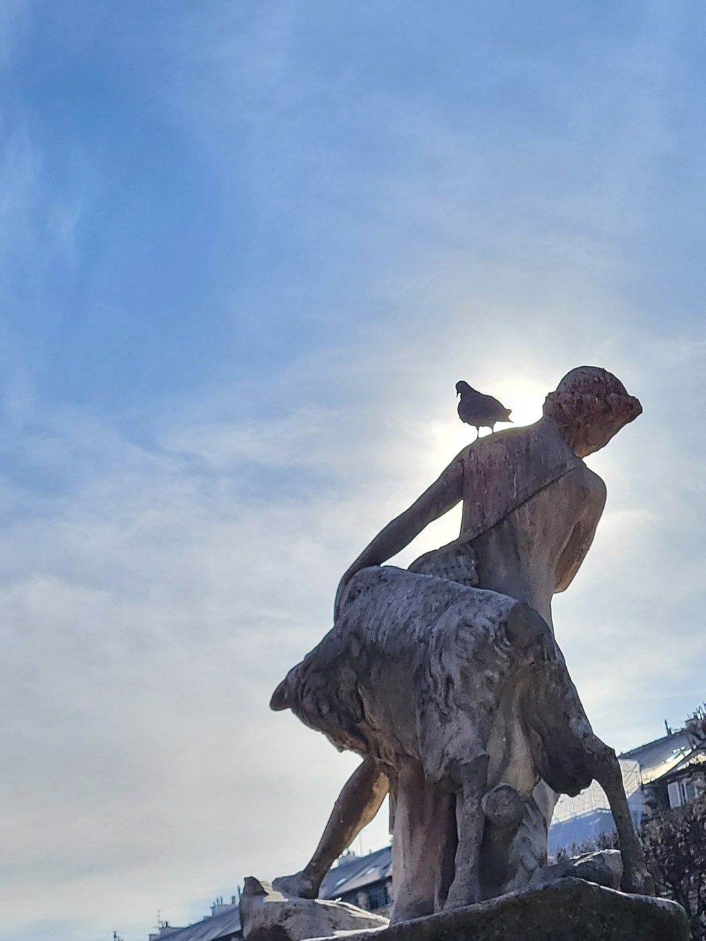 En prenant le soleil sur un banc : contre-jour au pigeon posé sur l&rsquo;épaule de la statue d&rsquo;un petit&nbsp;pâtre.