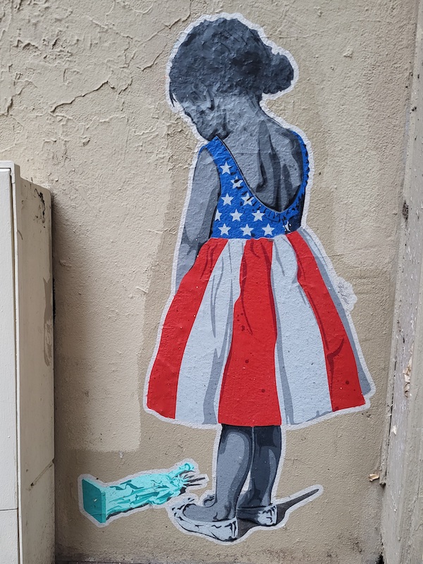 Au bas d&rsquo;un mur lézardé dans une rue de la ville, cette image : une petite fille de dos, vêtue d&rsquo;une jolie robe taillée dans un drapeau américain, regarde tristement son  jouet brisé, une statue de la liberté. (Paris&nbsp;2026)